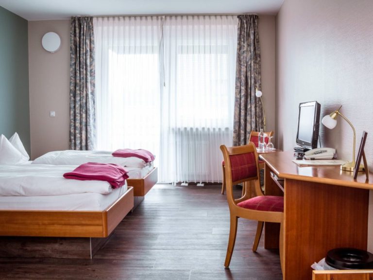 Ein großes, helles, gemütlich eingerichtetes Zweibettzimmer mit einem großen Fenster und einem Schreibtisch.