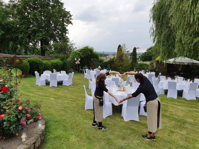 Garten mit festlich gedeckten Tischen und zwei Servicekräfte, die aus ihren Armen ein Herz formen.