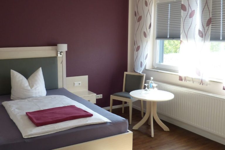 Hotelzimmer mit Einzelbett, Tisch und Stuhl, bordeauxroter Wand und hellem Fenster.