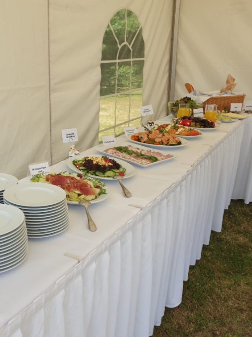 Ein Buffet mit verschiedenen Gerichten auf einem langen Tisch - gedeckt mit weißen Skirtings und weißen Tellern - in einem Zelt im Garten.