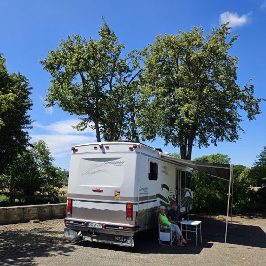 Ein Wohnmobil parkt auf einem Parkplatz, umgeben von Bäumen und blauem Himmel.