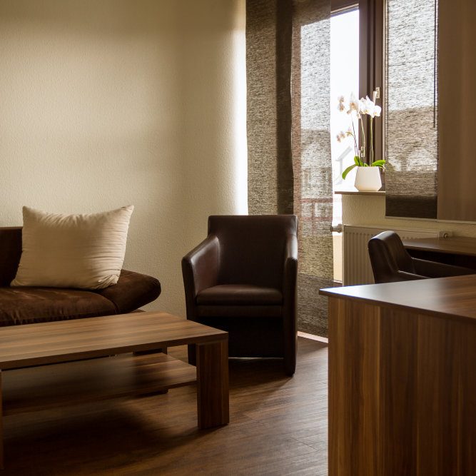 Ein gemütliches Zimmer mit Couch, Sessel, Schreibtisch und Schreibtischstuhl, in warmen Brauntönen eingerichtet, mit Blick auf einen Balkon.